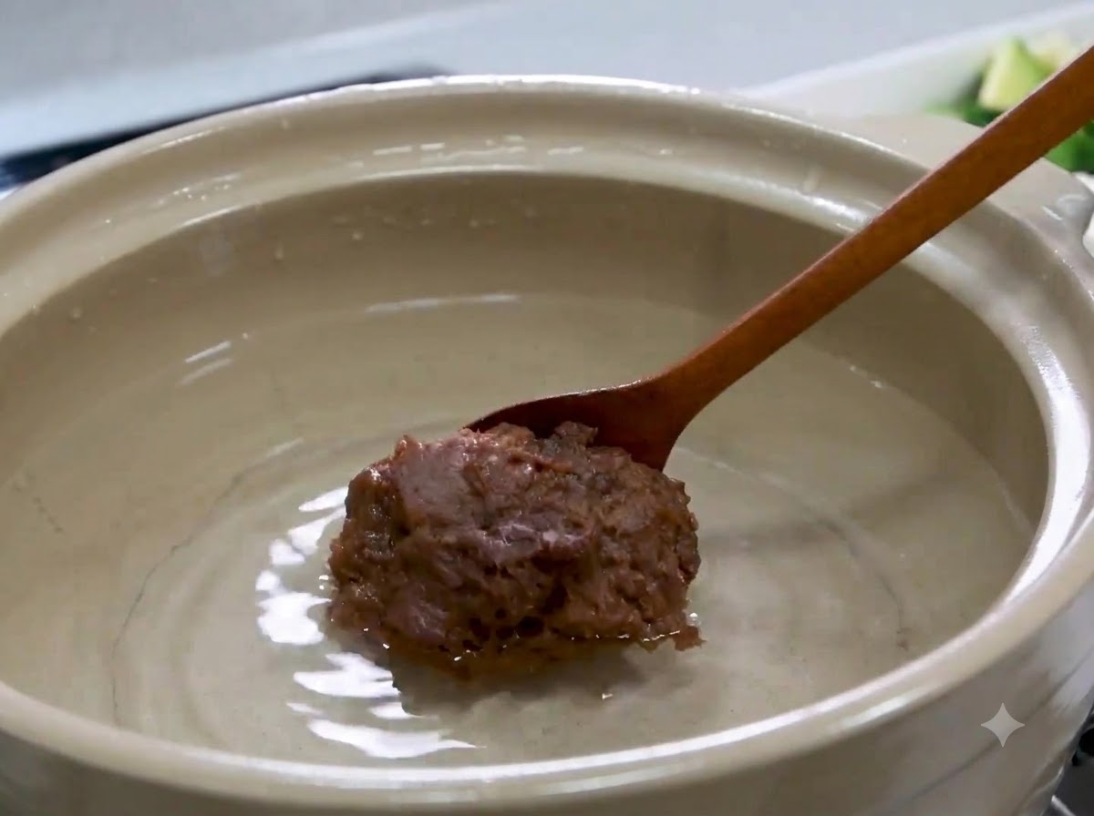 Una cuchara de madera sosteniendo una gran porción de pasta de soja coreana sobre el agua dentro de una olla.