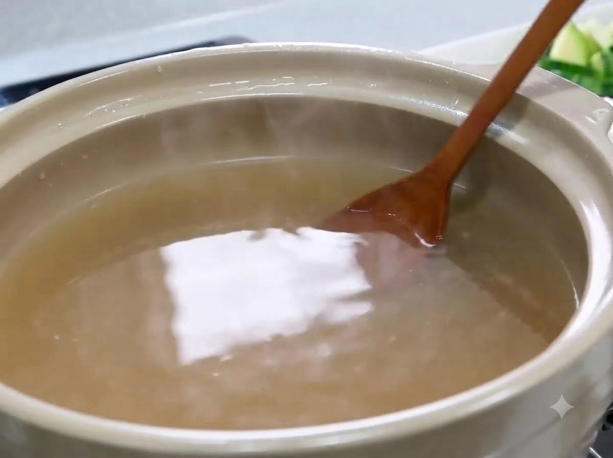 Una cuchara de madera removiendo y disolviendo pasta de soja dentro de una olla con caldo marrón claro.