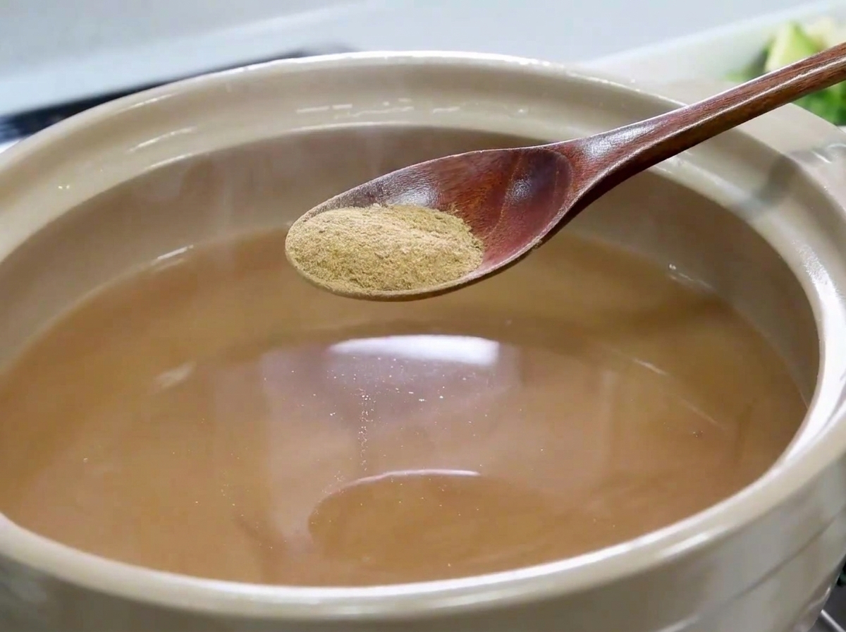 Una cuchara de madera sosteniendo polvo de anchoa fino sobre una olla llena de caldo marrón claro.