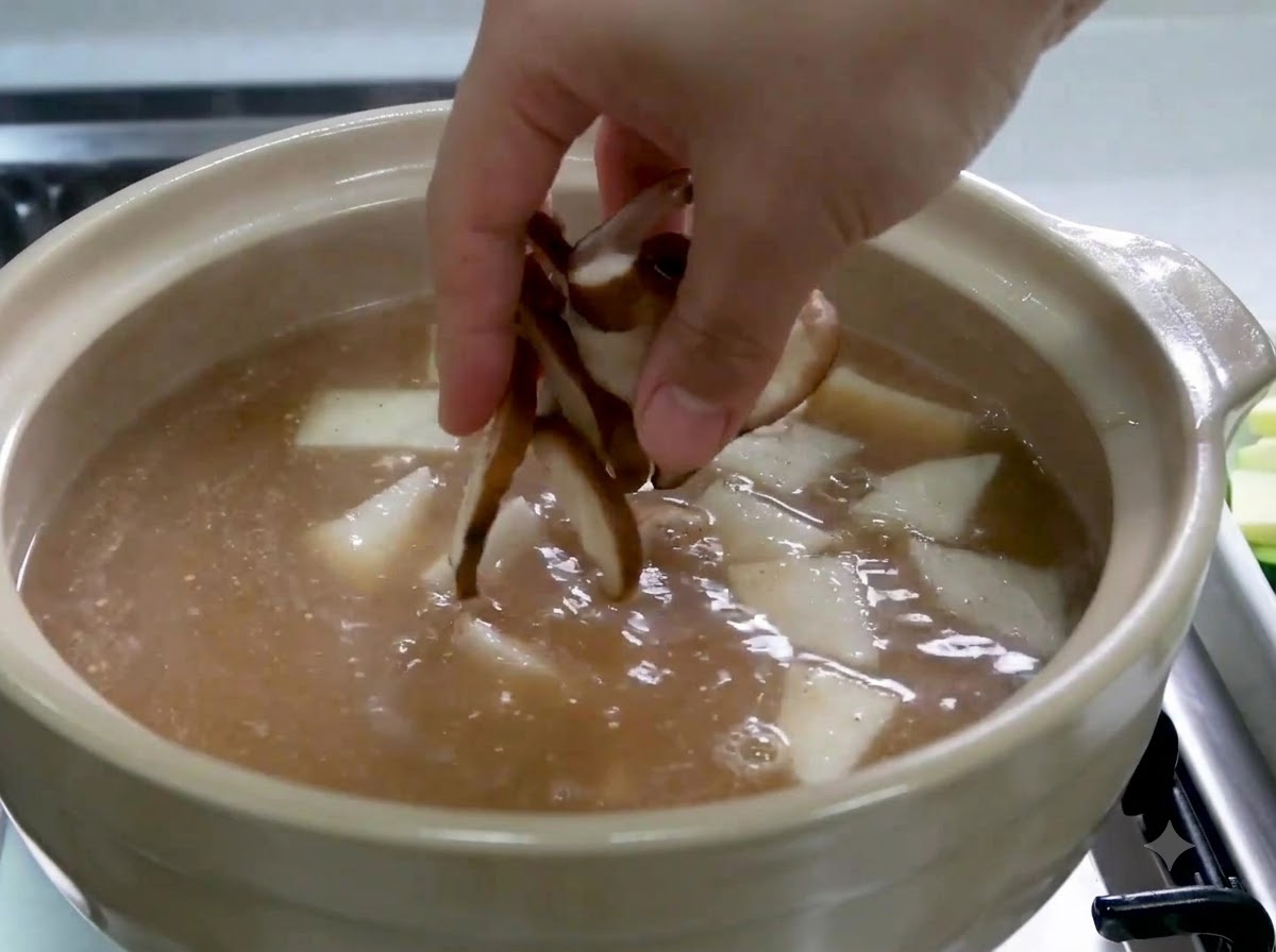Una mano echando setas shiitake en rodajas en una olla que contiene caldo y rodajas de rábano.