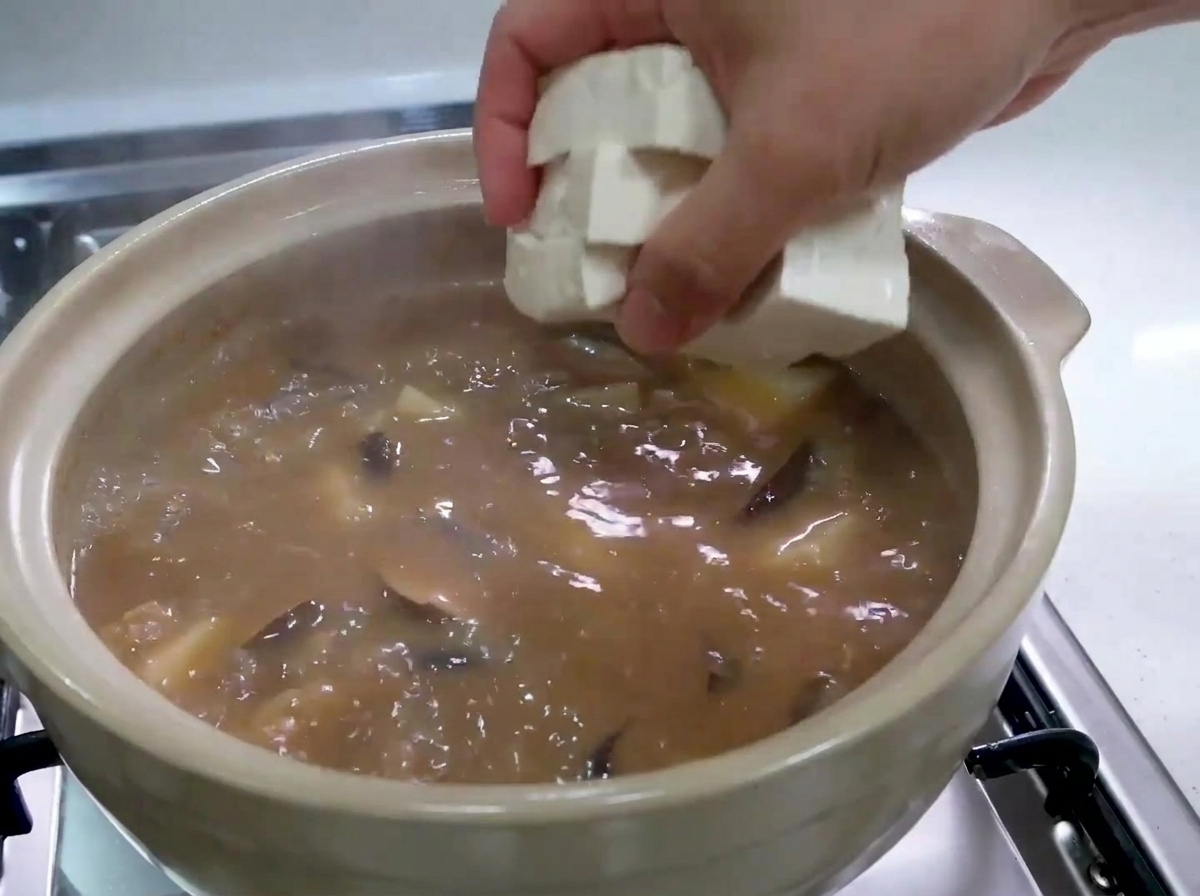 Una mano dejando caer con cuidado cubos de tofu blanco y fresco en una olla de estofado marrón burbujeante.