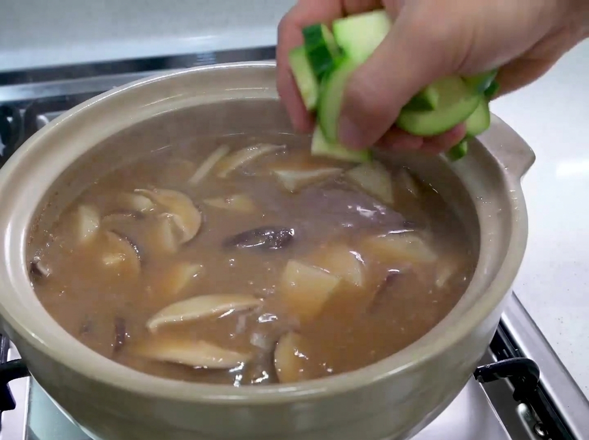Una mano echando trozos cortados de calabacín verde crudo en un estofado coreano burbujeante.