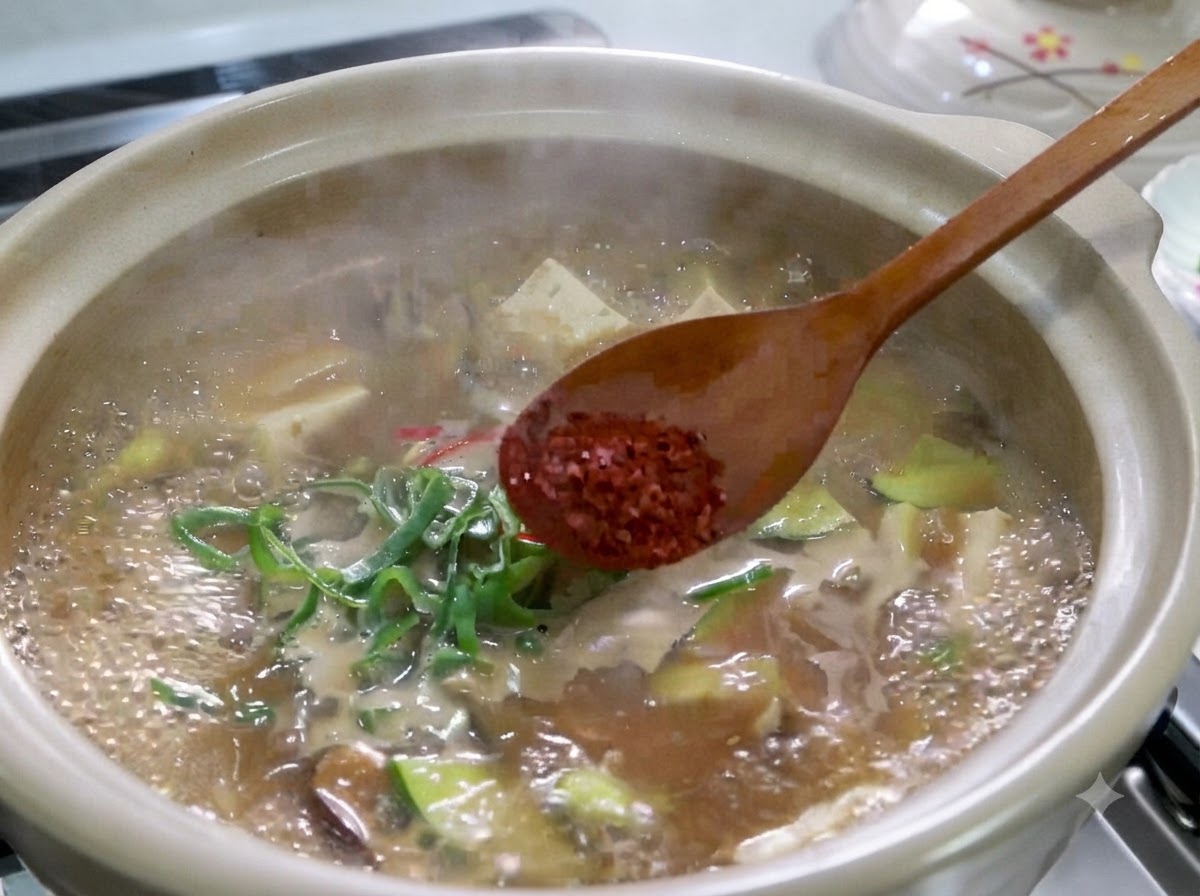 Una cuchara de madera sosteniendo chile rojo en polvo sobre una olla hirviendo de estofado de pasta de soja coreana.