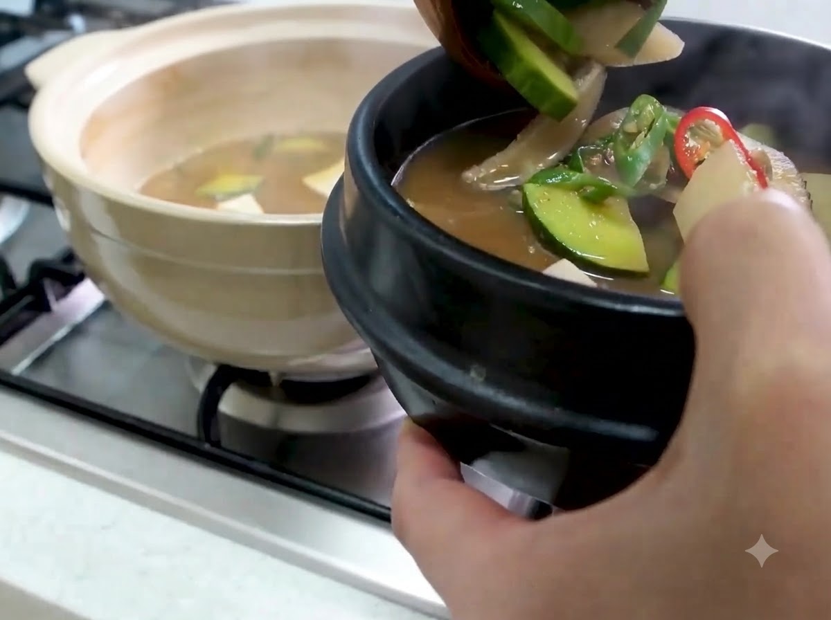 Un cazo transfiriendo Doenjang Jjigae caliente con calabacín y verduras a un cuenco de piedra negra para servir.