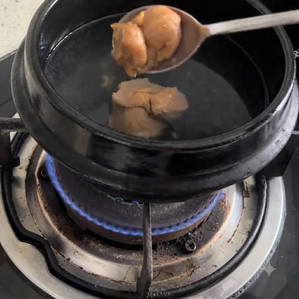 Una cuchara con pasta de soja siendo vertida en una olla negra con agua hirviendo sobre una estufa.