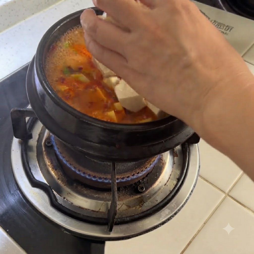 Cubos de tofu blanco siendo añadidos a mano en una olla de barro con sopa picante.