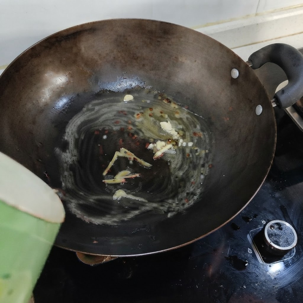 Agua hirviendo siendo vertida desde un recipiente verde en un wok que contiene manteca derretida, jengibre y ajo.