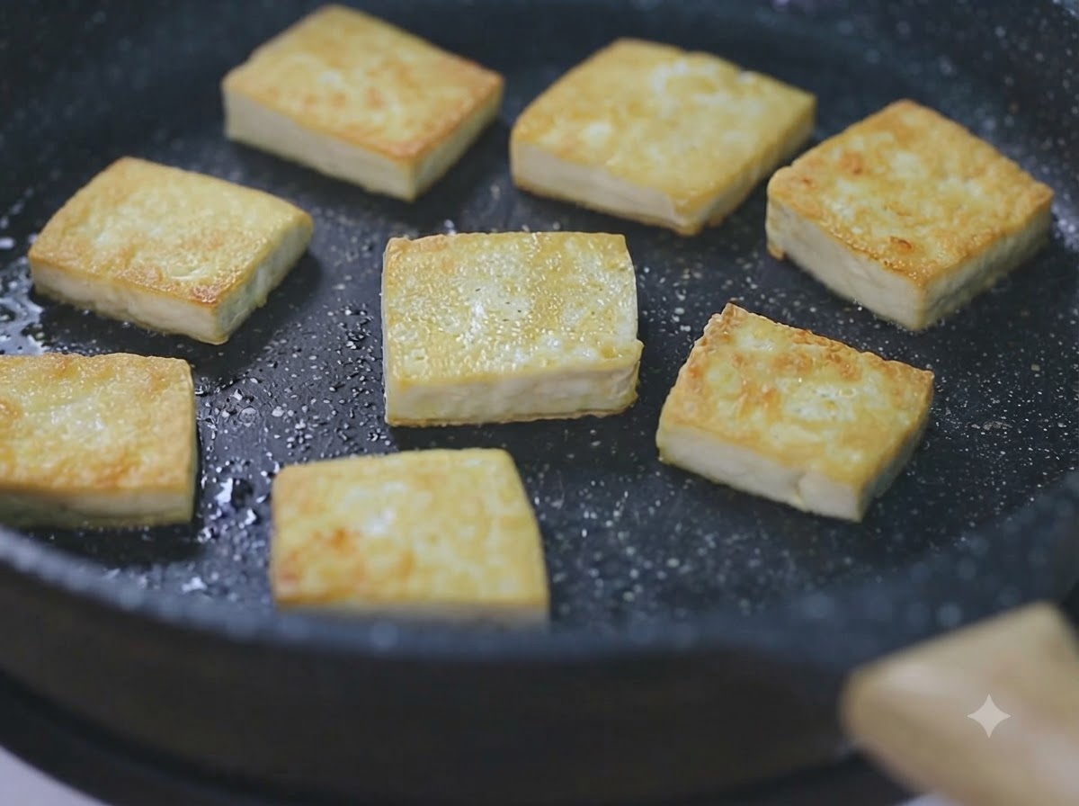 Varios cubos de tofu dorados friéndose en una sartén negra a fuego lento.