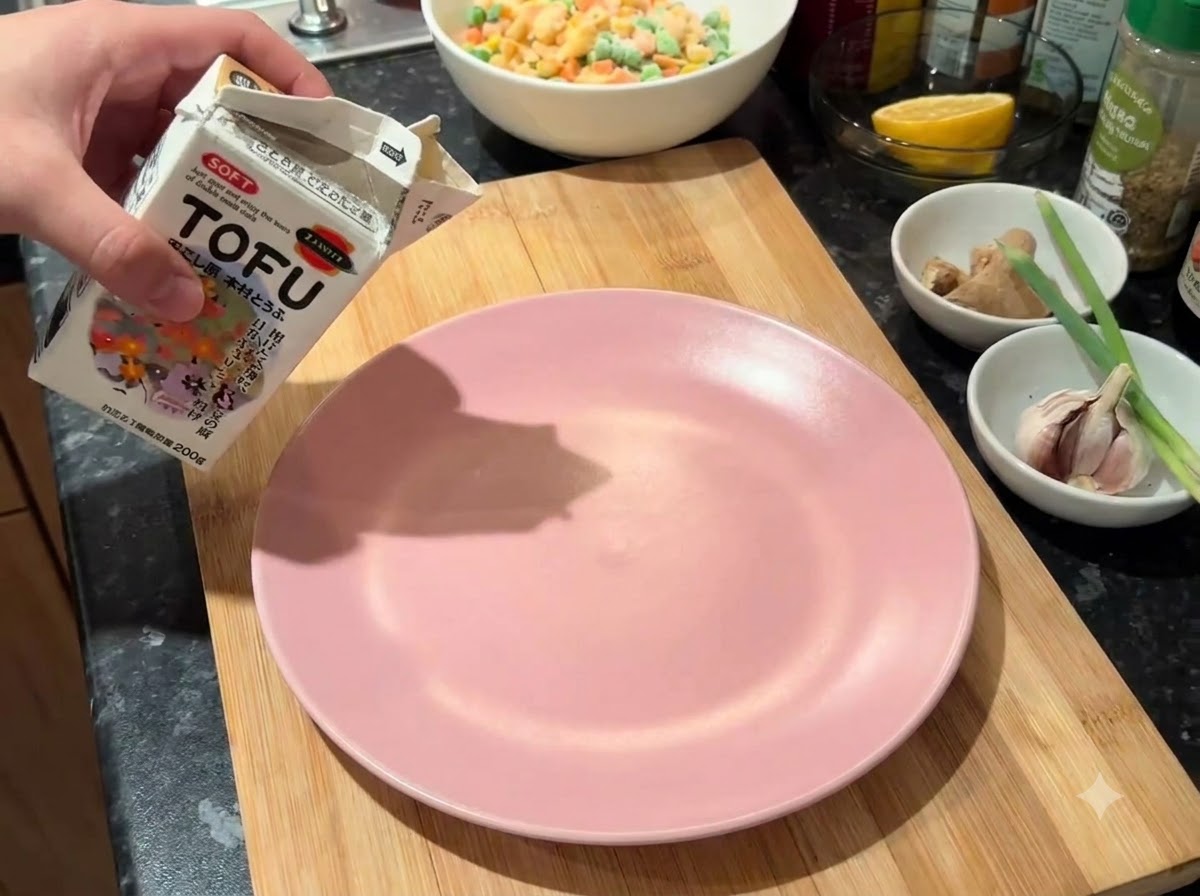 Un cartón de tofu blando siendo vaciado sobre un plato rosado en una tabla de cortar de madera.