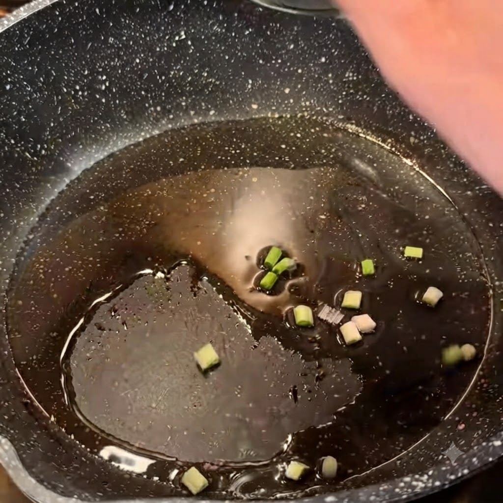 Cebollas verdes picadas chisporroteando en aceite caliente dentro de una sartén oscura.