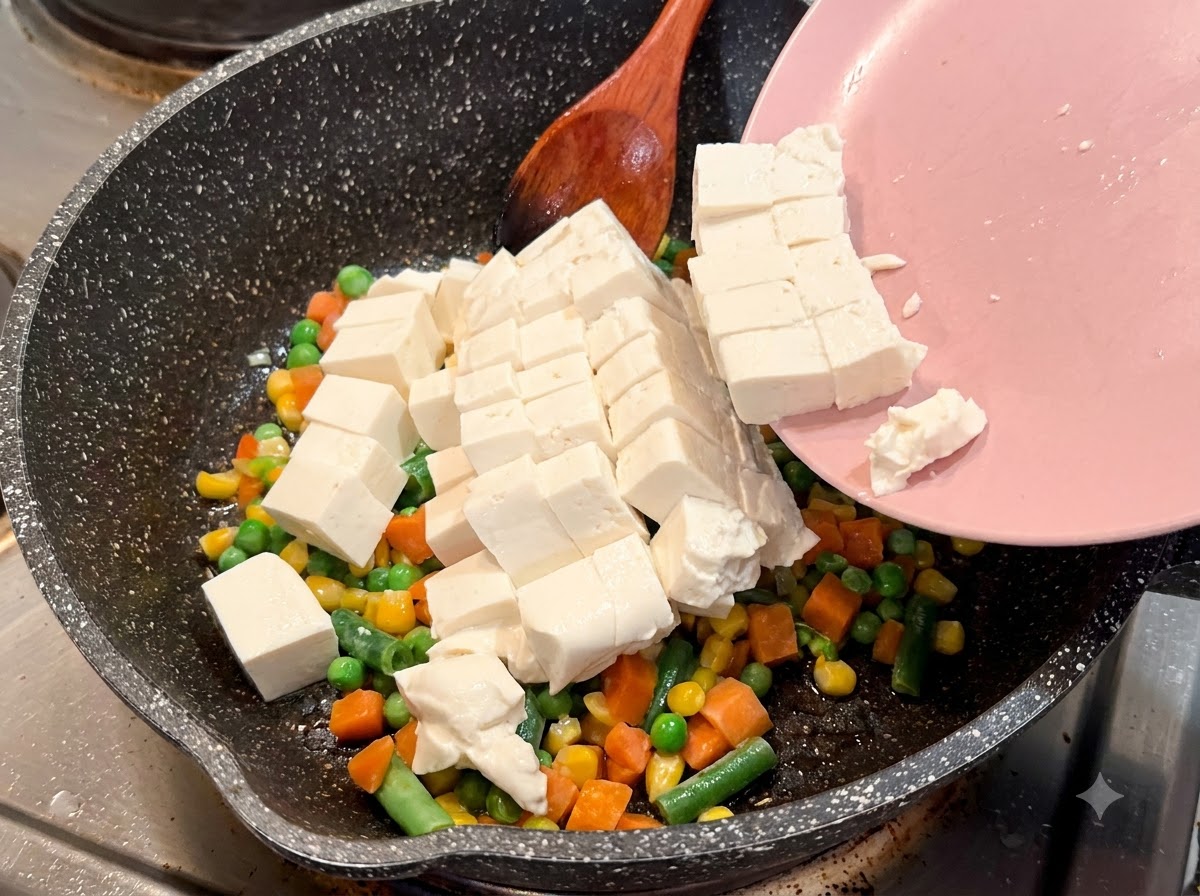 Tofu blando en cubos deslizándose de un plato rosado a una sartén moteada con verduras mixtas.