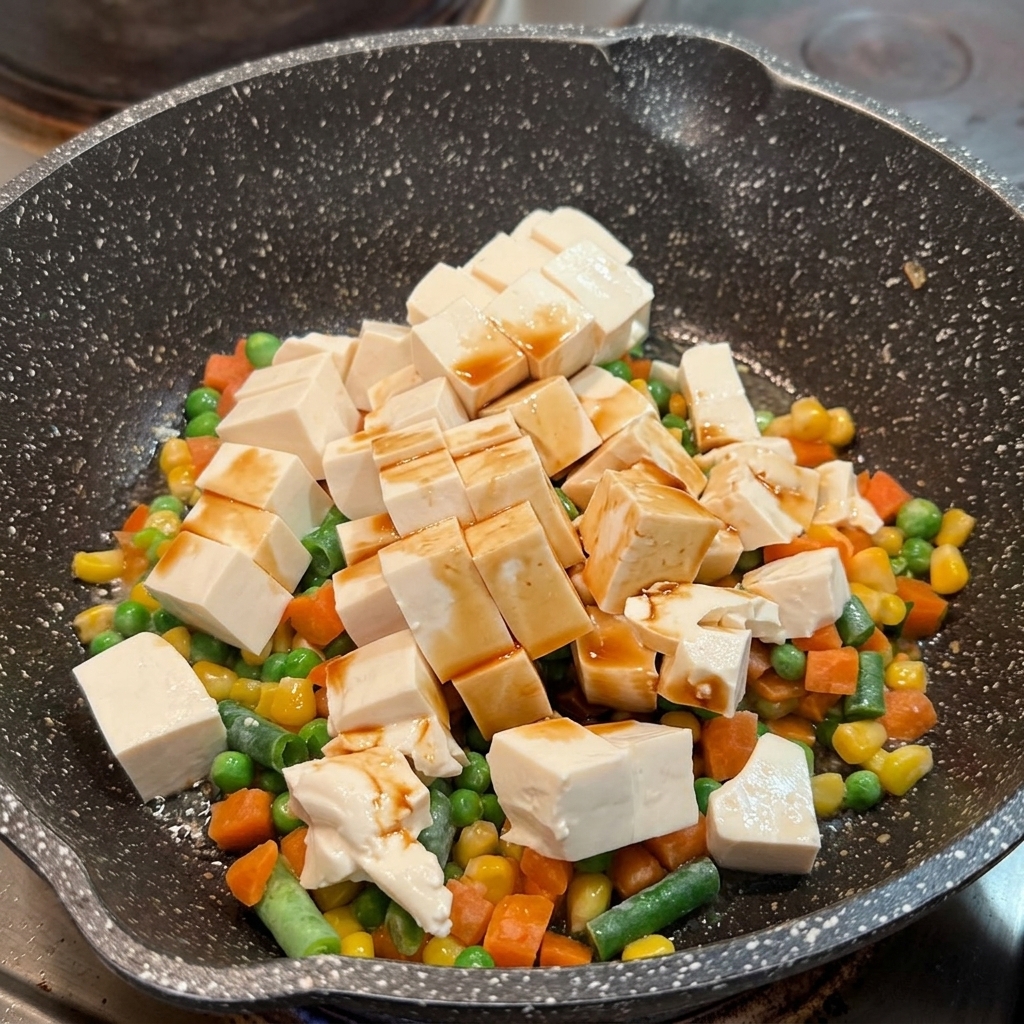 Salsa de soja distribuida sobre cubos de tofu blando y verduras mixtas en una sartén.