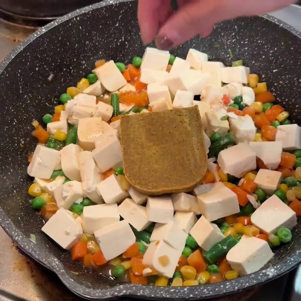 Una mano colocando un bloque de roux de curry marrón sólido sobre el tofu y las verduras en una sartén.
