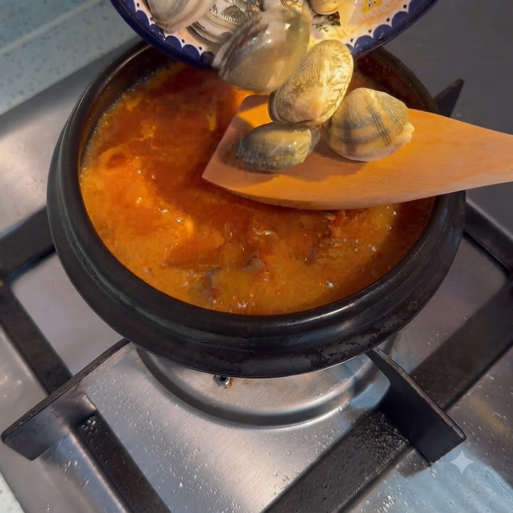 Almejas frescas vertiéndose desde un plato pequeño a una olla burbujeante de estofado coreano.