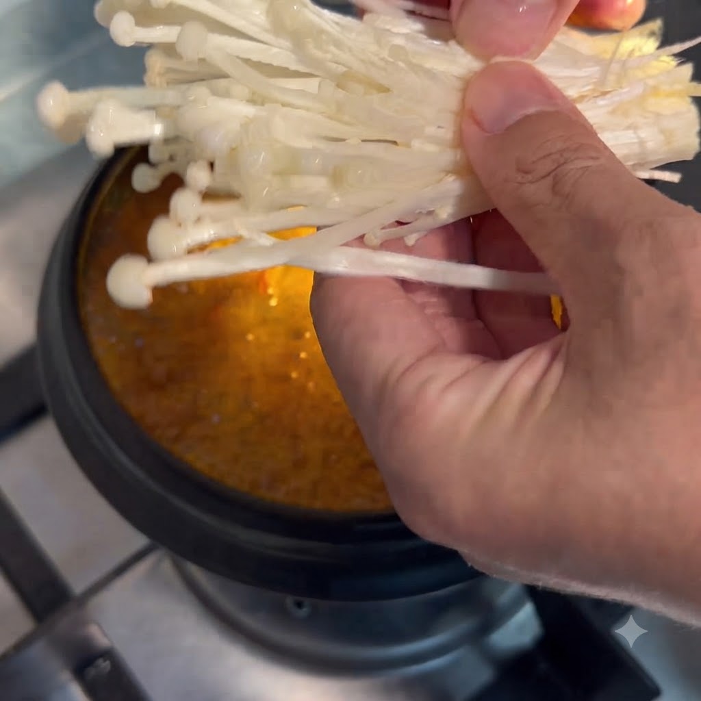 Hongos enoki sostenidos con la mano y siendo bajados a la olla de sopa a fuego lento.
