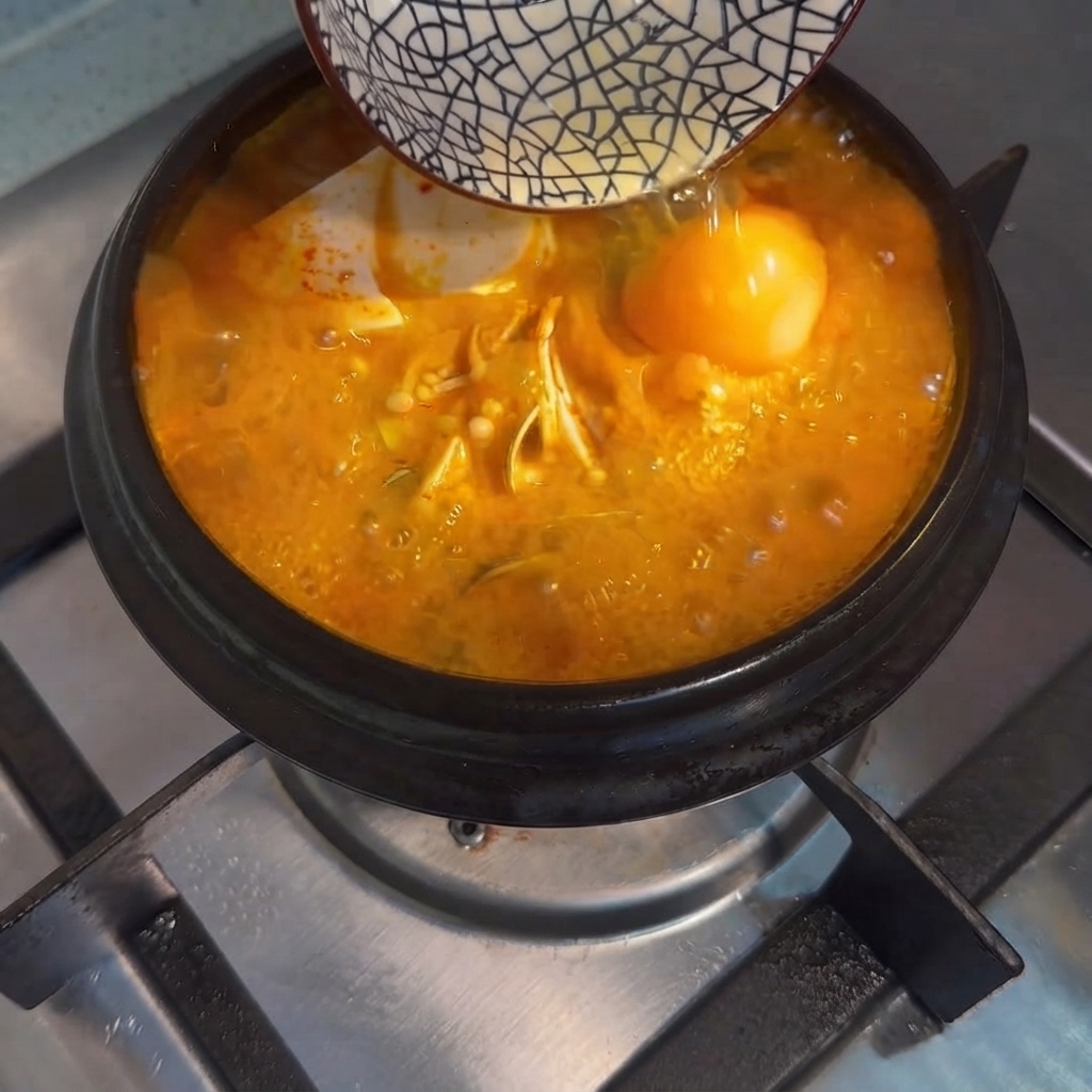 Un huevo crudo vertiéndose desde un cuenco pequeño en el estofado de tofu coreano burbujeante.