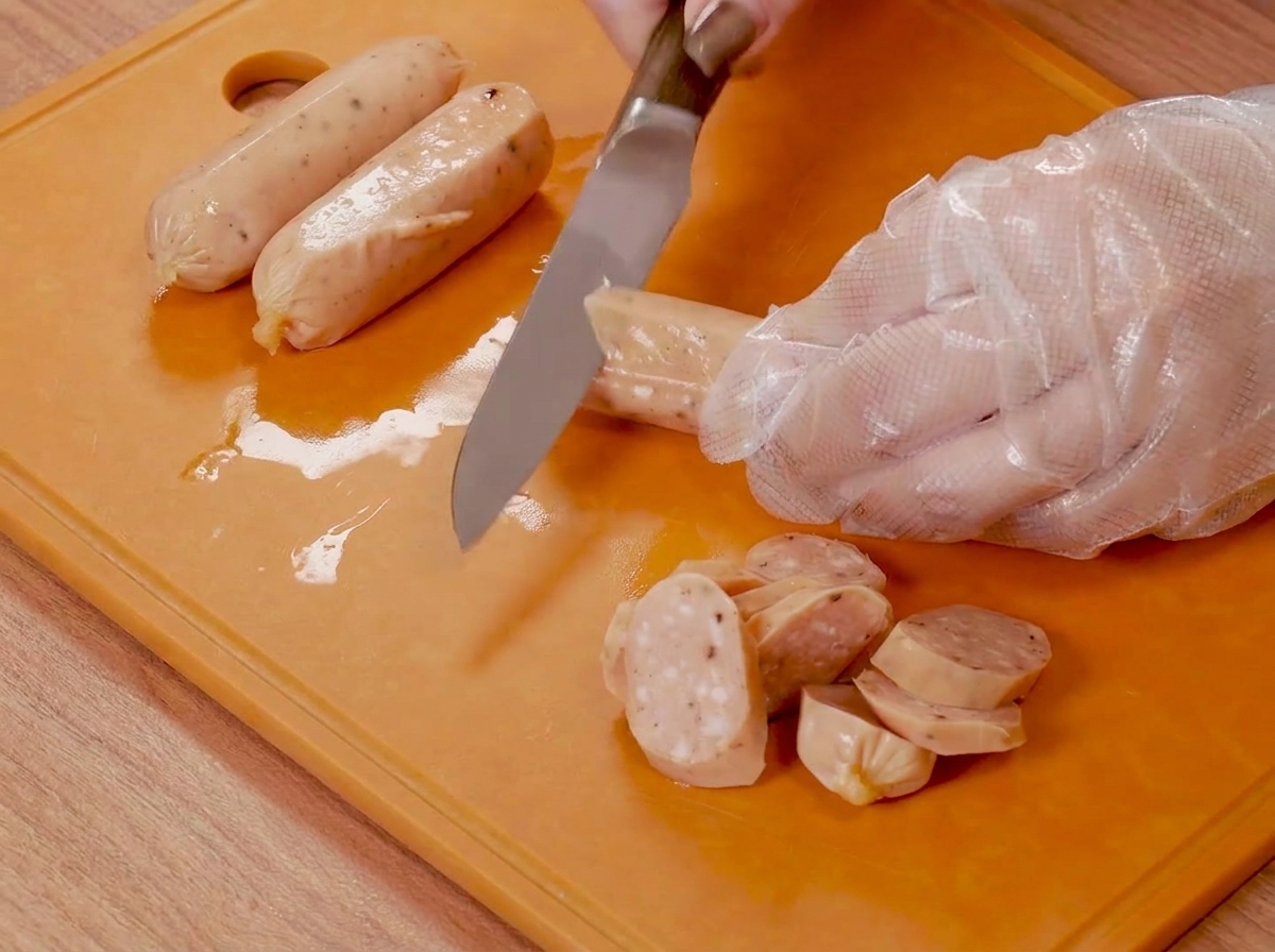 Manos con guantes de plástico usando un cuchillo para hacer cortes en salchichas pequeñas sobre una tabla de cortar naranja.