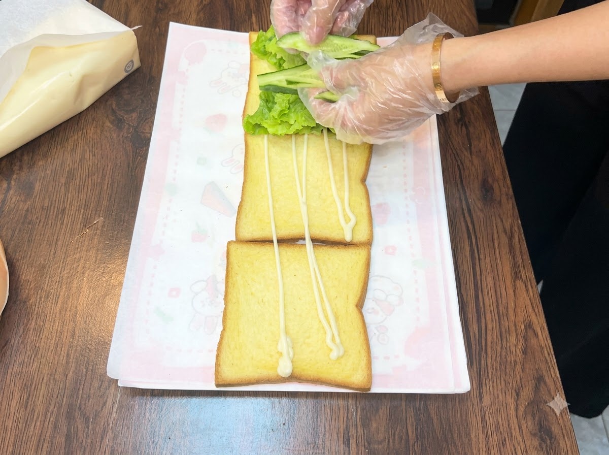 Manos acomodando rodajas de pepino verde fresco sobre una cama de lechuga en un trozo de pan tostado.