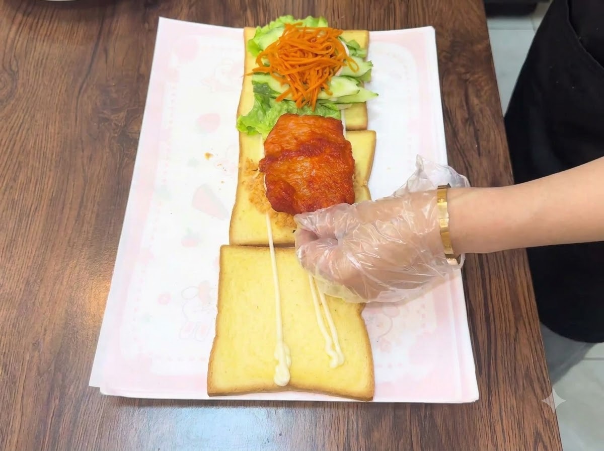 Una mano con guante colocando una milanesa de pollo frita y caliente sobre el ensamblaje de pan tostado en capas.