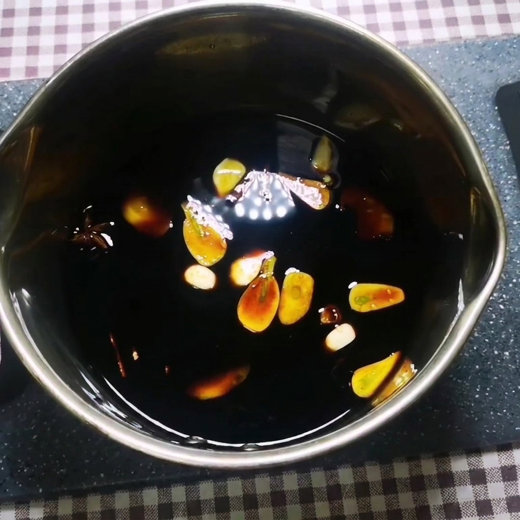 Una salsa oscura hirviendo a fuego lento en una olla de metal con ajo, jengibre, anís estrellado y canela flotando en su interior.