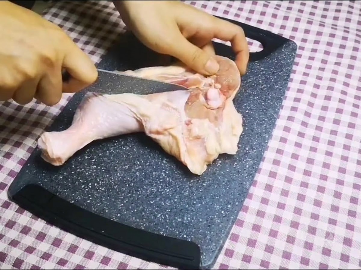 Un cuchillo afilado cortando a lo largo del hueso de un muslo de pollo crudo en una tabla de cortar para retirar el hueso.