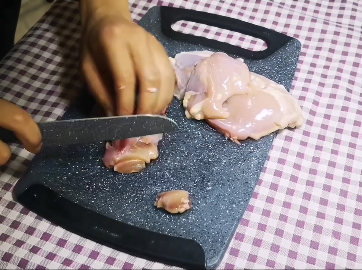 Un cuchillo cortando pollo crudo sin hueso en trozos pequeños sobre una tabla de cortar gris.