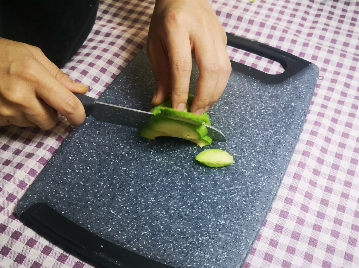 Manos cortando la mitad de un aguacate verde en trozos finos con un cuchillo sobre una tabla de cortar.