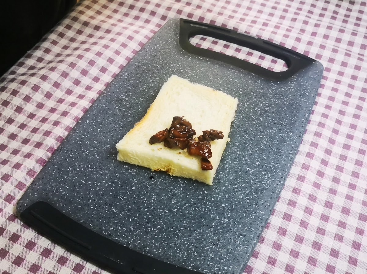Un primer plano de trozos de pollo glaseado oscuro colocados ordenadamente sobre una sola rebanada de pan de molde blanco que descansa sobre una tabla de cortar gris.