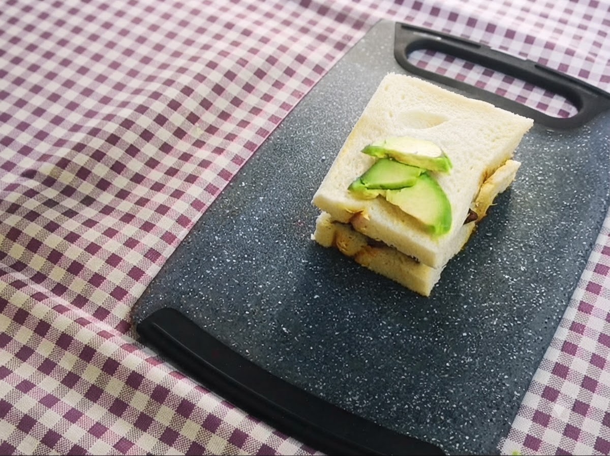Un sándwich en capas con aguacate en rodajas colocado encima, reposando sobre una tabla de cortar.