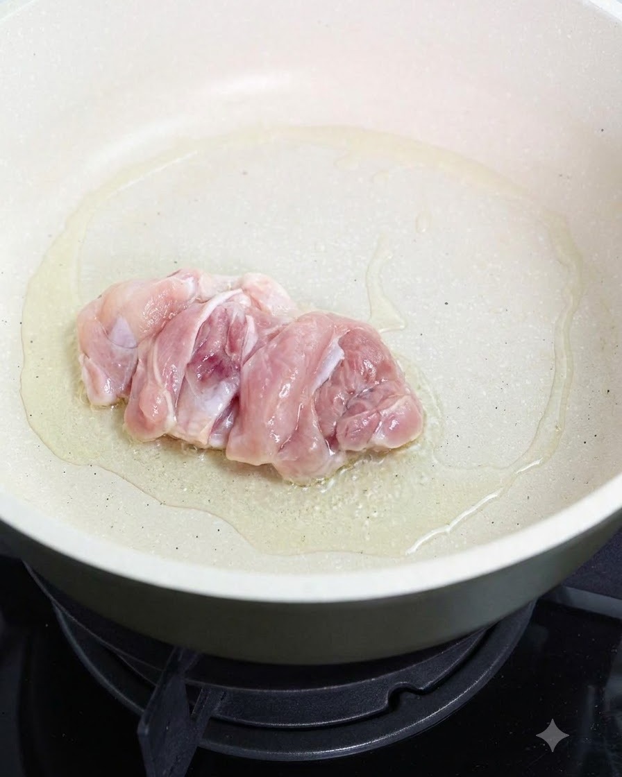Un muslo de pollo crudo friéndose con la piel hacia abajo en una sartén con aceite.