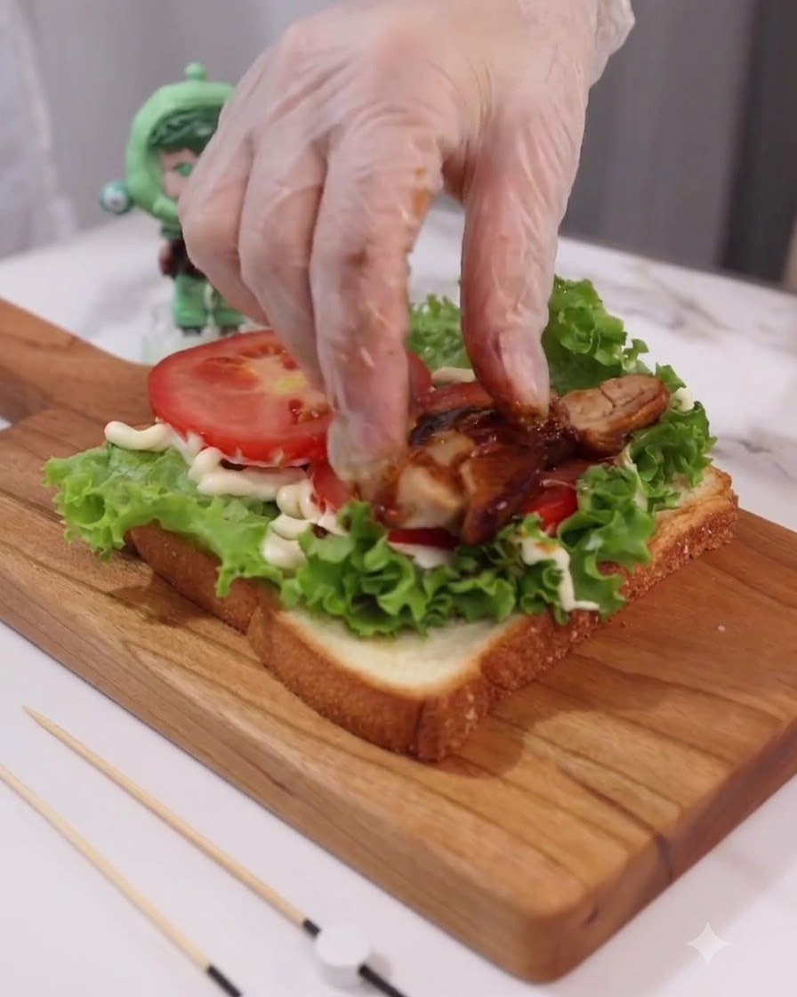Mano con guante colocando rodajas de pollo teriyaki cocido sobre las capas de tomate y lechuga.
