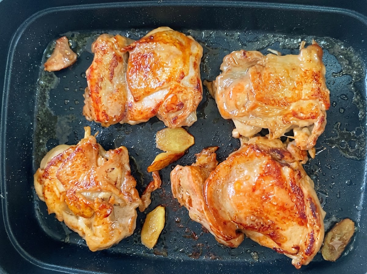 Cuatro muslos de pollo friéndose en una sartén con la piel crujiente y dorada junto a rodajas de jengibre.