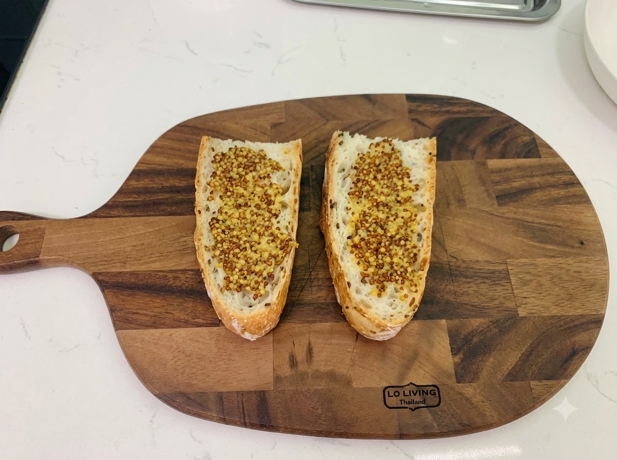 Dos rebanadas de pan rústico colocadas sobre una tabla de cortar de madera, ambas untadas uniformemente con mostaza en grano.
