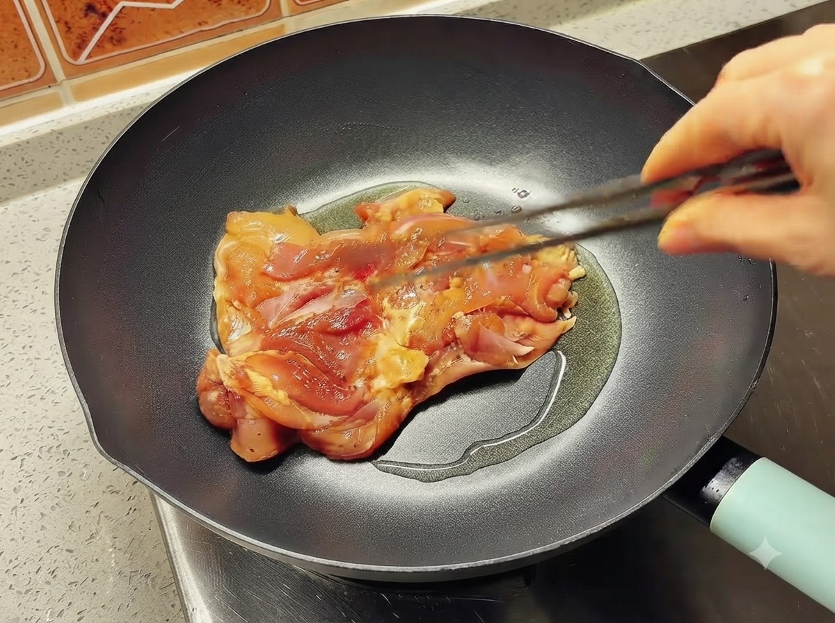 Una sola pieza de muslo de pollo marinada colocándose en una sartén caliente con aceite usando pinzas.