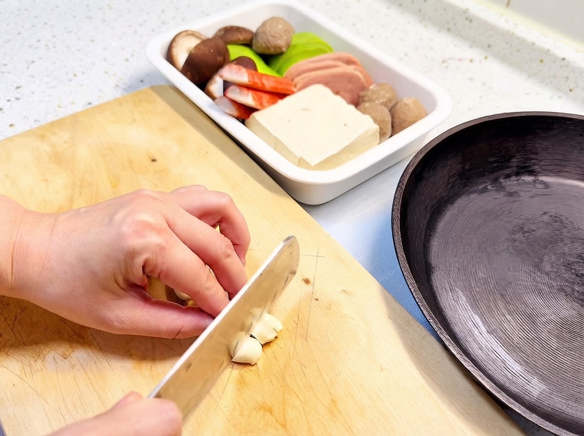 Dientes de ajo cortándose en una tabla de madera junto a una sartén negra y una bandeja con ingredientes.