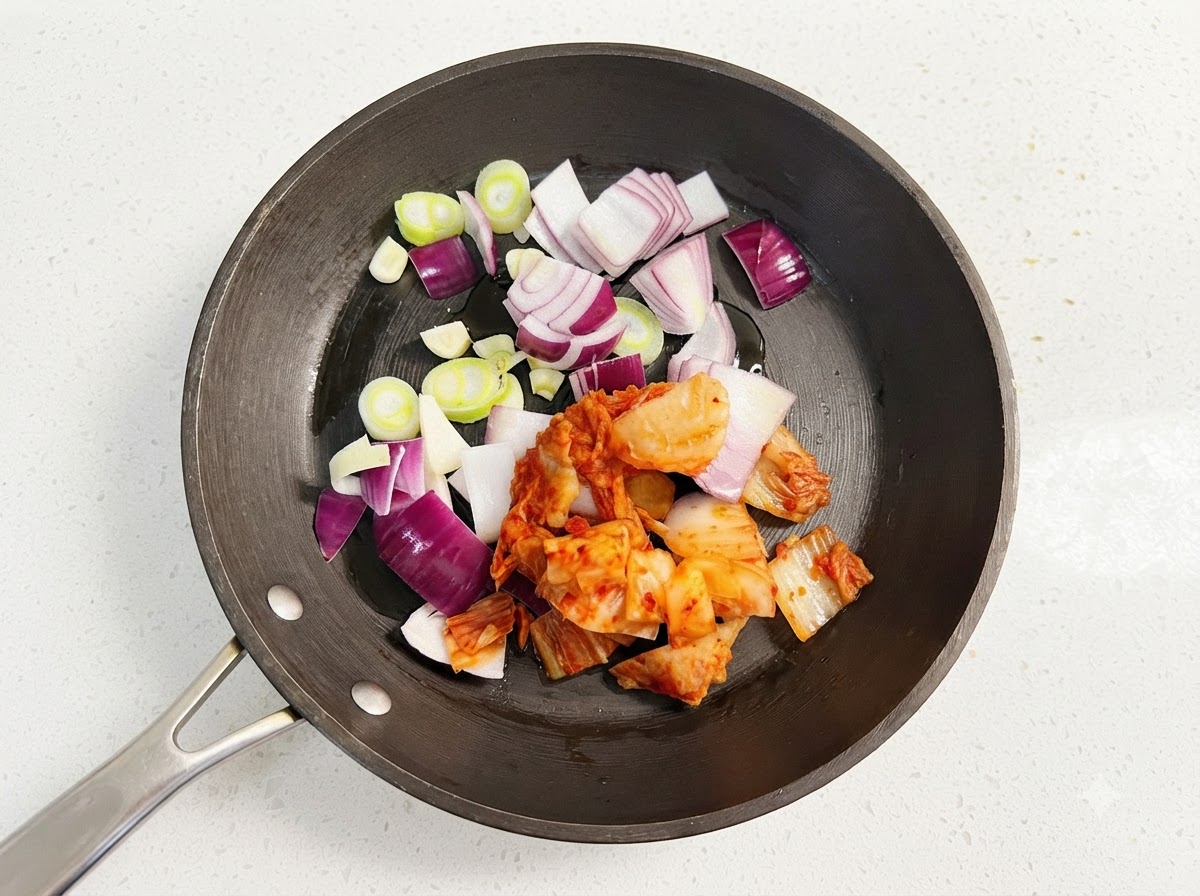 Tiras de cebolla morada, rodajas de cebolleta y kimchi rojo chisporroteando con aceite en una sartén.