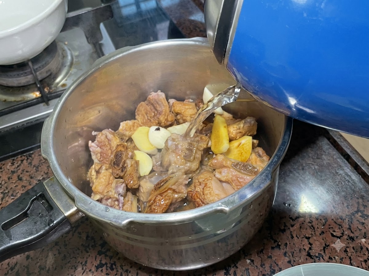 Agua siendo vertida desde una tetera eléctrica en una olla a presión de acero inoxidable llena de costillas sazonadas y aromáticos.