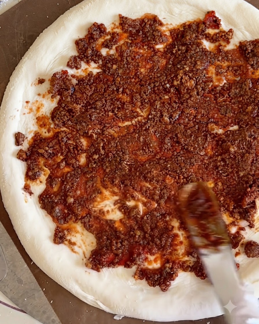 Dados de ternera cocidos esparcidos sobre la salsa de carne roja extendida uniformemente por la base de masa blanca de la pizza.