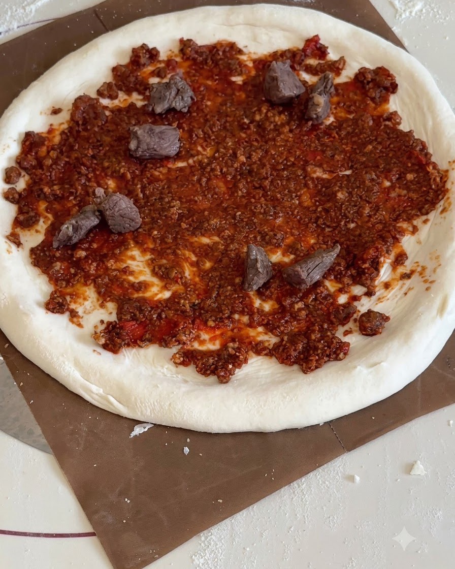 Trozos triangulares de piña amarilla y trozos de ternera cocida distribuidos sobre la base de pizza con salsa roja.