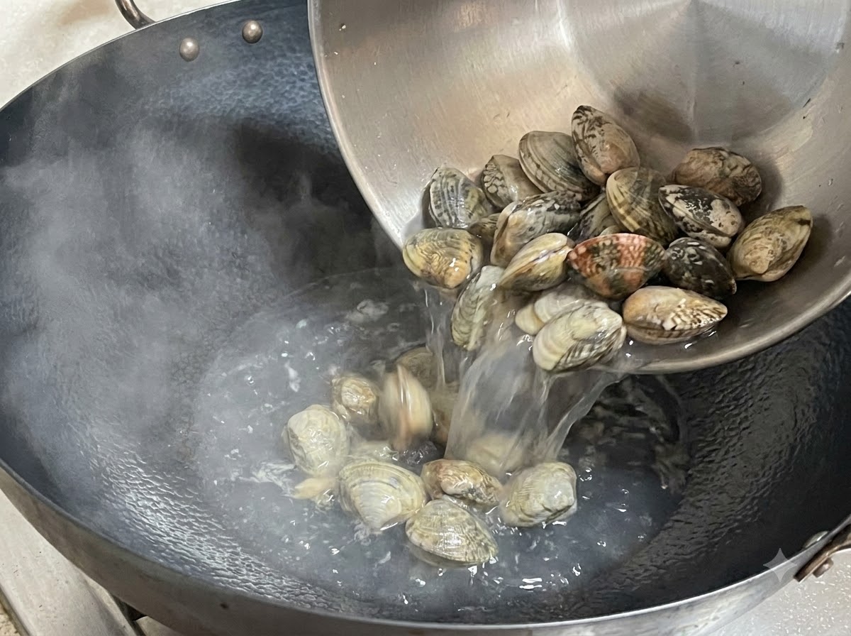 Almejas frescas siendo vertidas desde un bol de metal en un wok humeante.