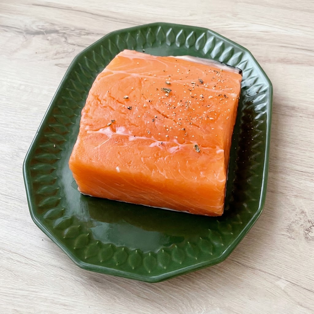 Un trozo grueso de salmón fresco sobre un plato verde, generosamente espolvoreado con sal marina gruesa y pimienta negra molida.
