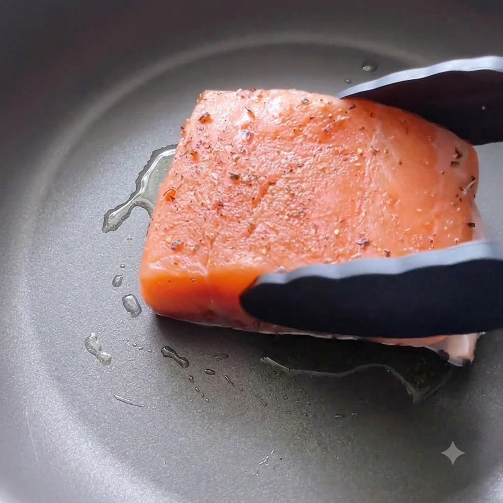 Un filete de salmón sazonado siendo bajado con la piel hacia abajo en una sartén con aceite usando pinzas de cocina negras.