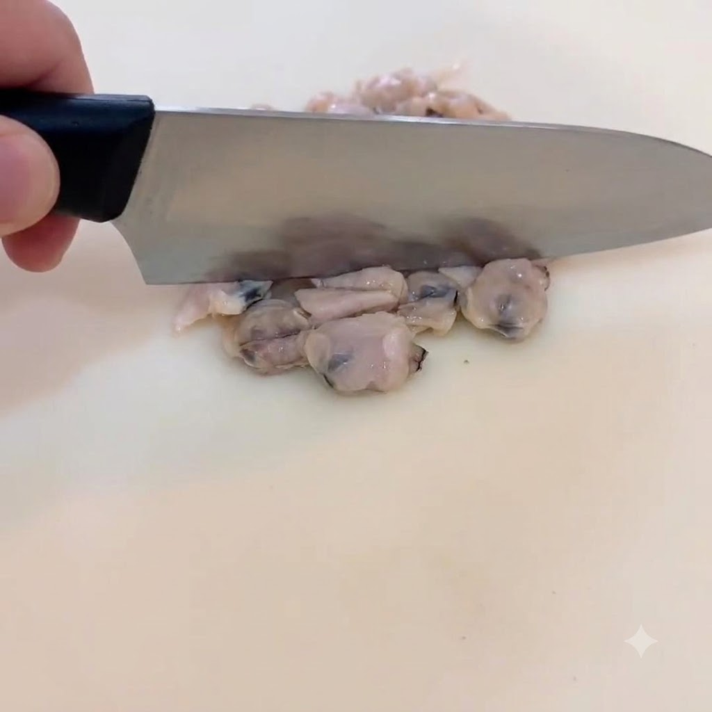 Un primer plano de una mano sosteniendo un cuchillo de chef, picando suavemente carne de almeja cocida en trozos pequeños sobre una tabla de cortar blanca.
