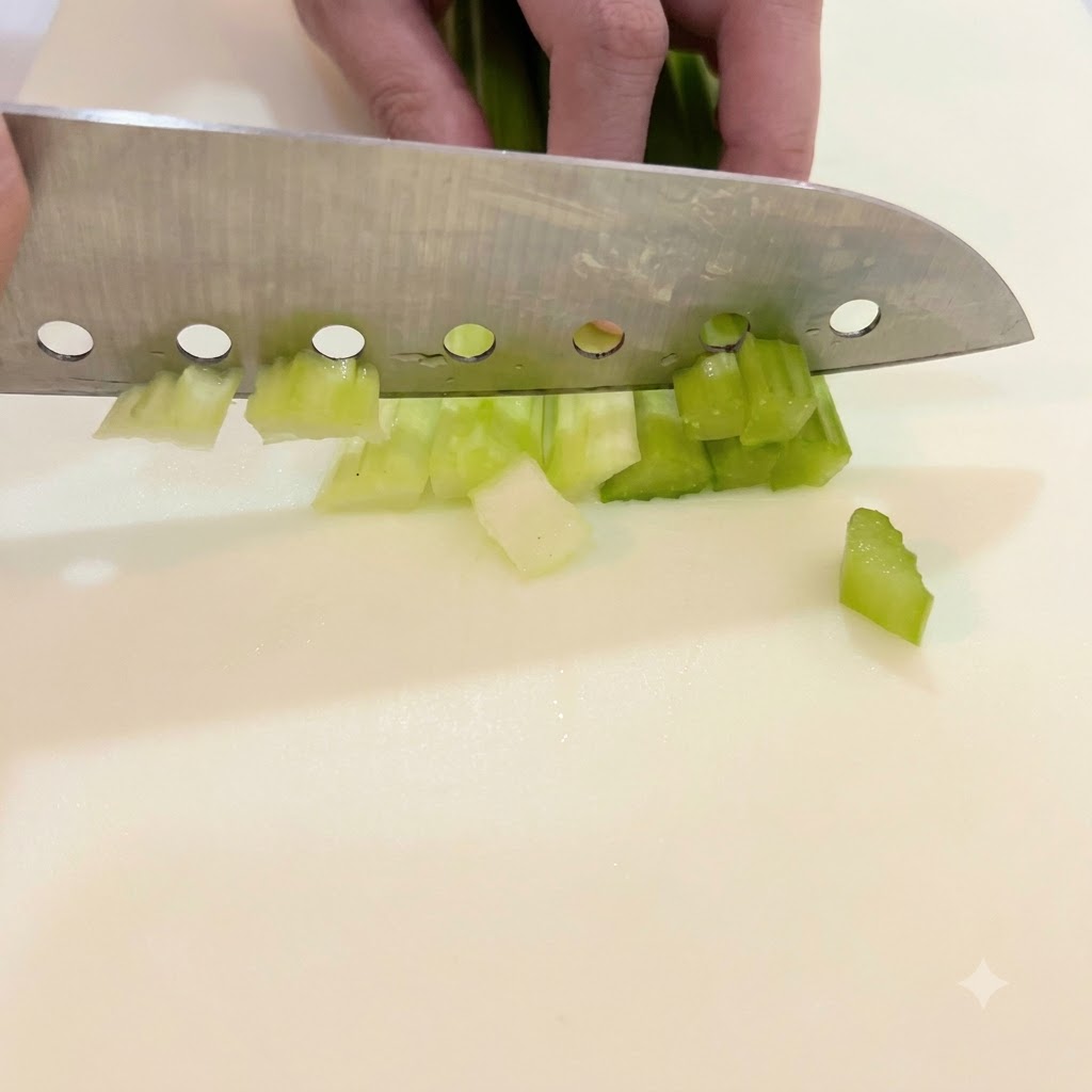 Una mano guiando un cuchillo grande para cortar tallos de apio verde brillante en trozos pequeños y uniformes sobre una tabla de cortar blanca.