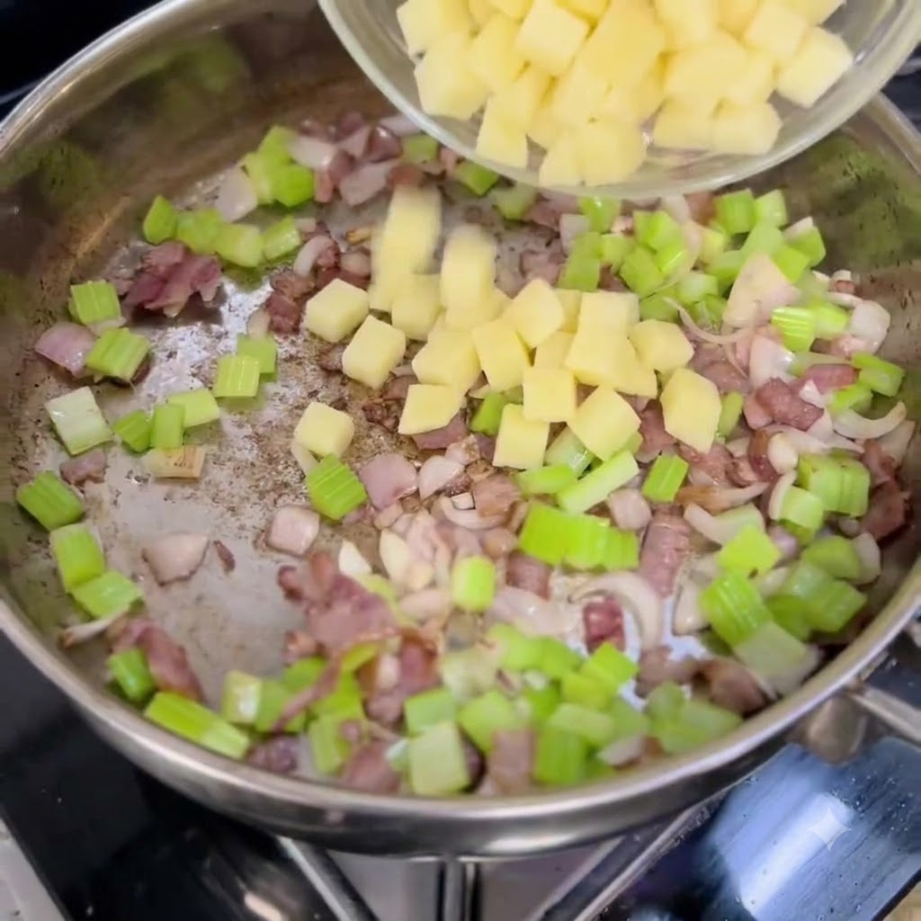 Patatas en dados siendo vertidas desde un tazón transparente en una sartén que contiene cebollas, apio y tocino picados.