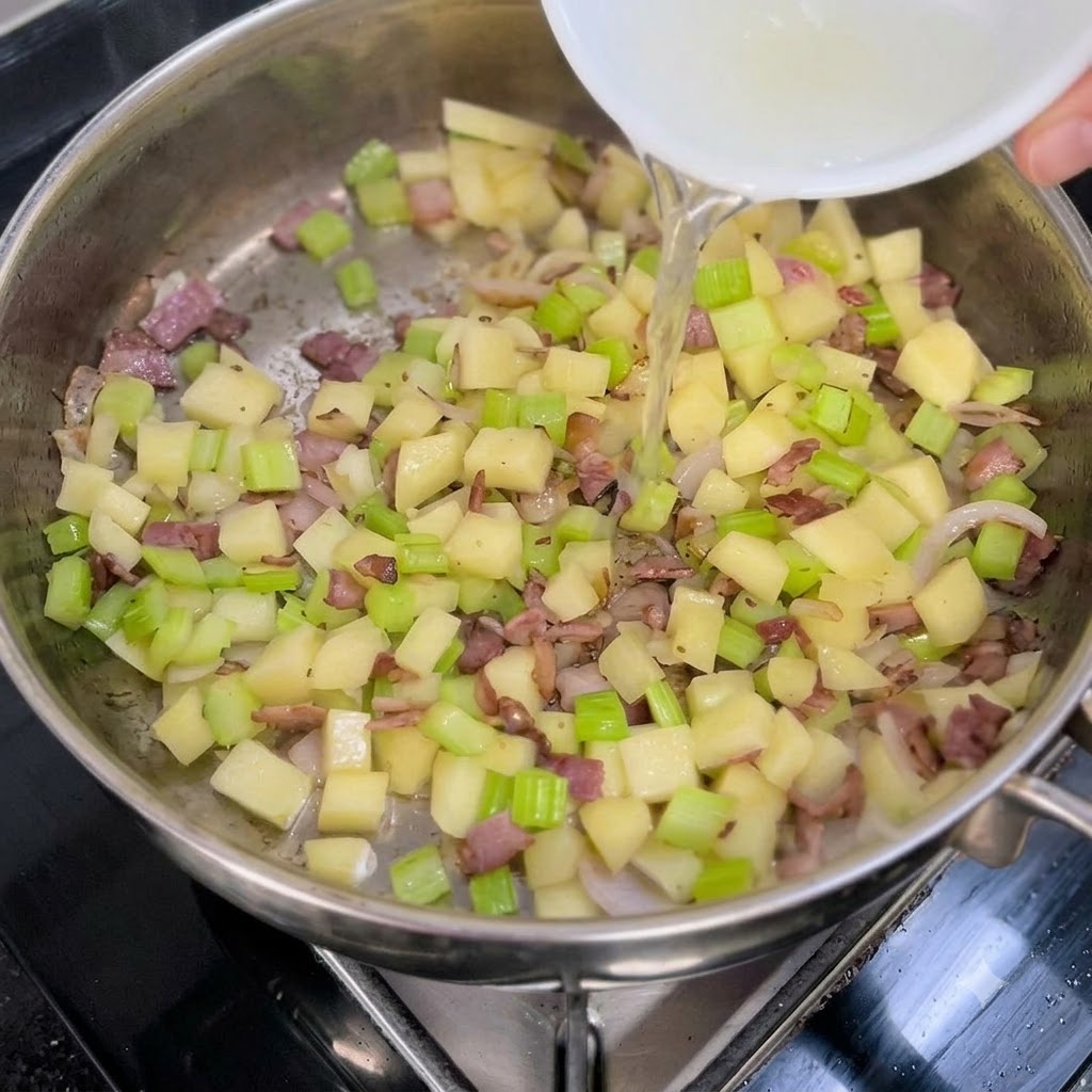 Caldo de almejas transparente siendo vertido desde un tazón blanco en una sartén con verduras salteadas y tocino.