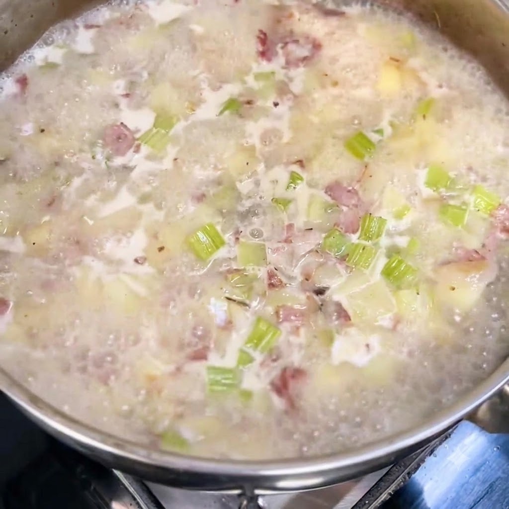 Leche y crema de leche siendo vertidas en una olla de sopa hirviendo a fuego lento con trozos visibles de patatas, apio y tocino.