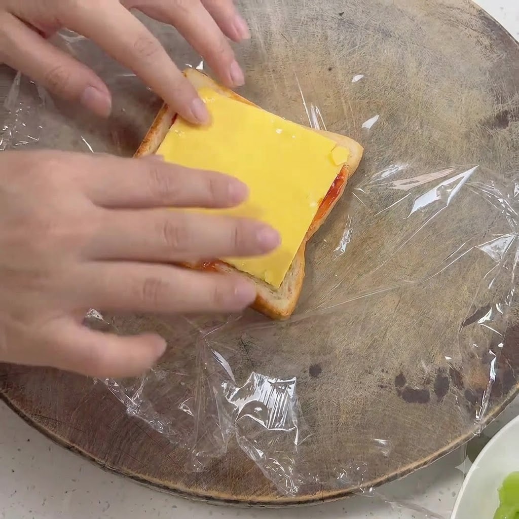 Manos colocando una rebanada de queso amarillo cuadrado sobre un trozo de pan cubierto con salsa.