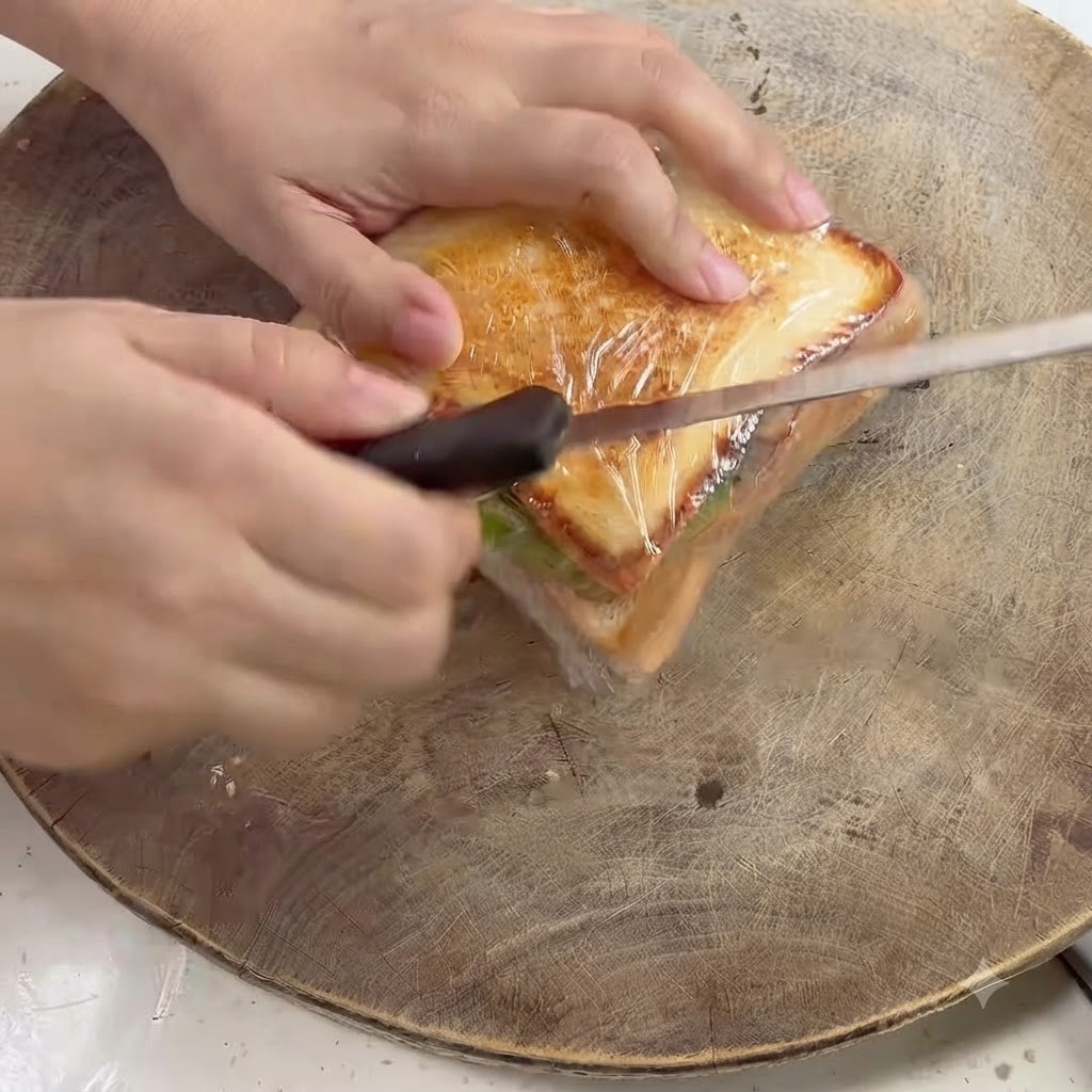 Un cuchillo de cocina cortando diagonalmente a través de un sándwich envuelto en plástico sobre una tabla de madera.