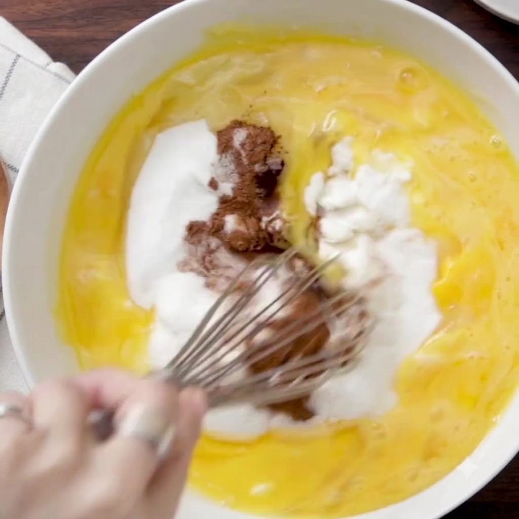Un batidor de alambre mezclando ingredientes húmedos, azúcar y canela dentro de un tazón blanco grande.
